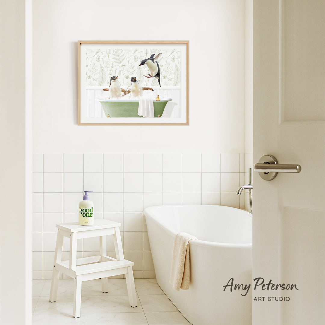 A framed painting of penguins sitting in a bathtub hangs on a white tiled wall above a white bathtub in a bathroom.