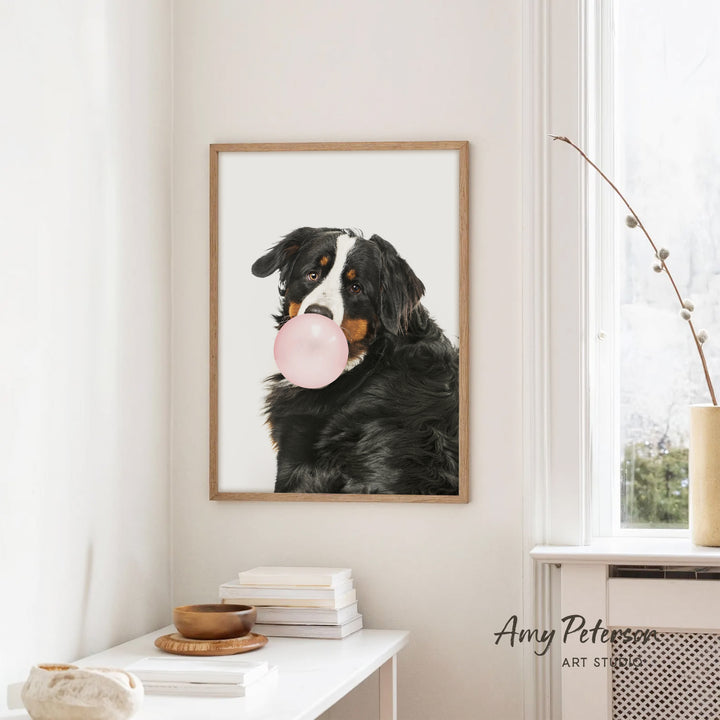 A framed image of a black and white dog blowing a bubble with a pink bubble gum bubble, hanging on a white wall in a room with a white table and a plant.