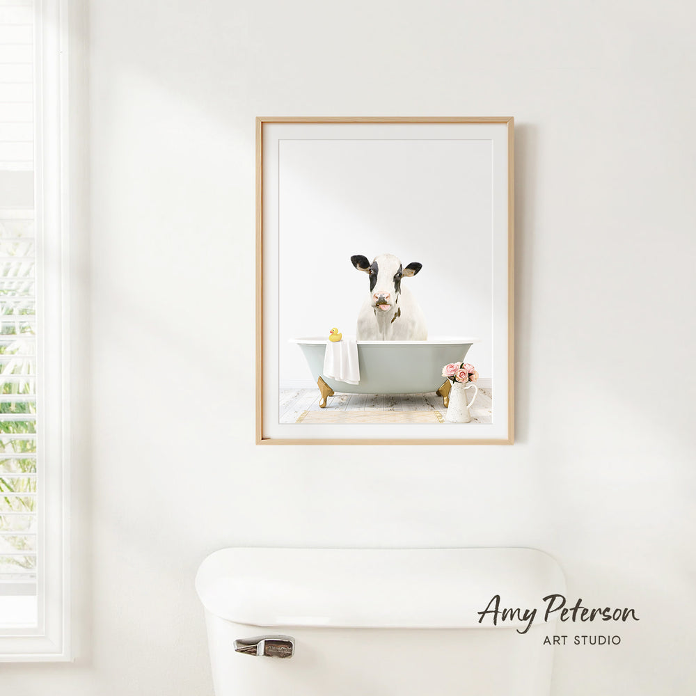 A framed image of a cow sitting in a bathtub, surrounded by flowers and a vase, is displayed on a white wall in a bathroom.