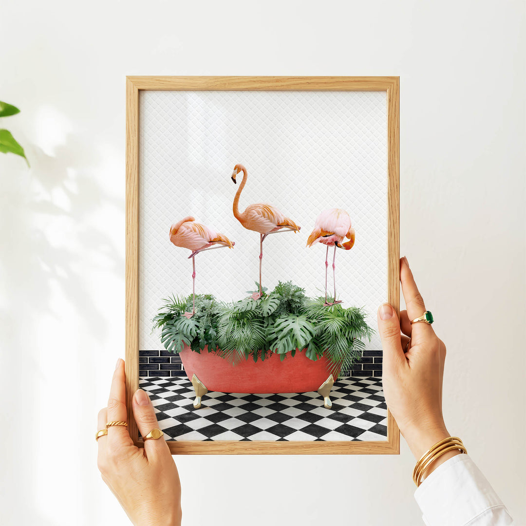 A person's hands are holding a framed picture of three flamingos standing in a red planter filled with green plants, set against a black and white checkered floor.