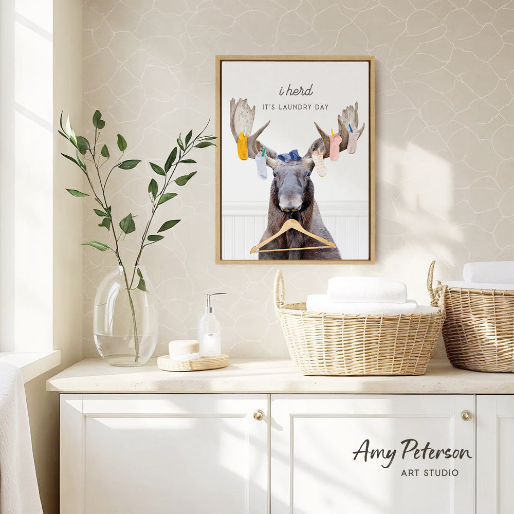 a framed poster of a moose with a humorous caption, hanging on a wall above a white cabinet with baskets and a plant.