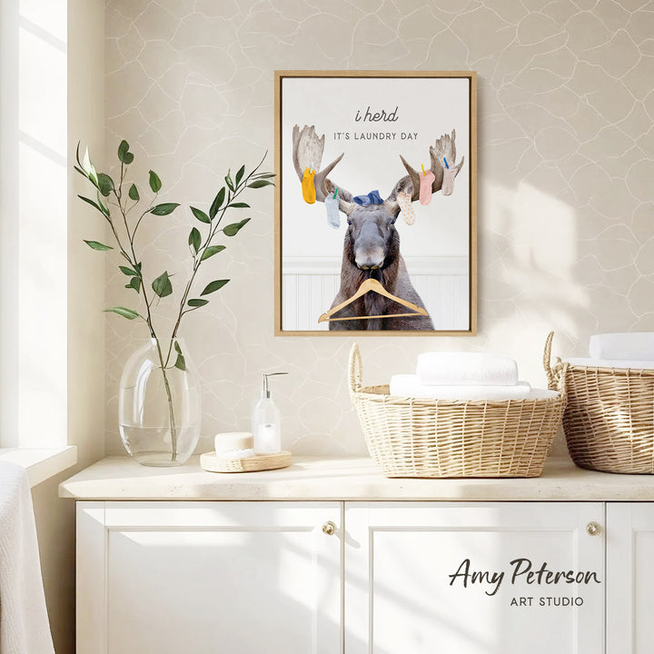 a framed poster of a moose with a humorous caption, hanging on a wall above a white cabinet with baskets and a plant.