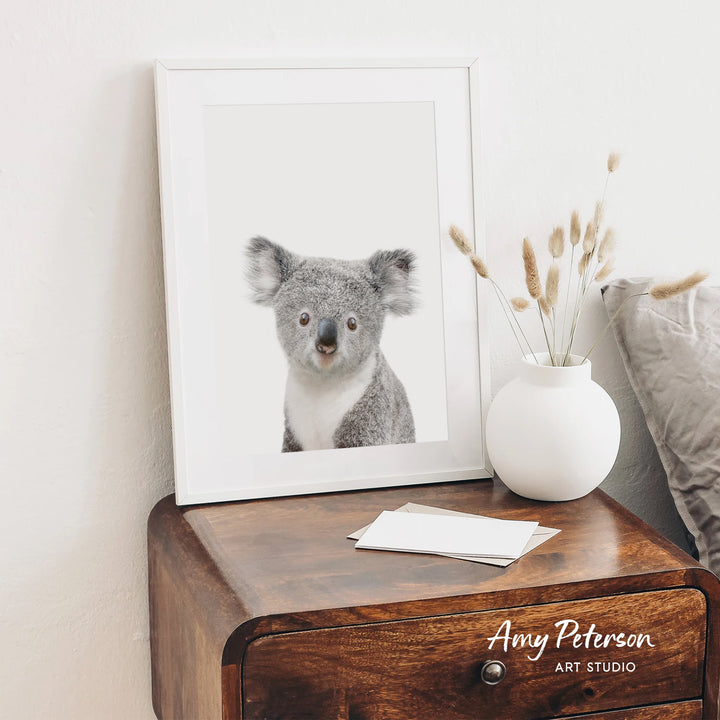 a koala bear in a white frame, placed on a wooden dresser next to a white vase with dried flowers.