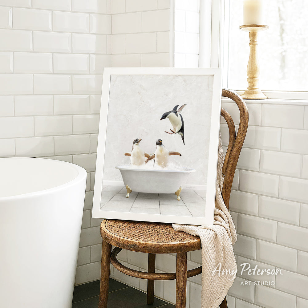 A framed image of penguins sitting in a bathtub, with one penguin standing on its hind legs, is displayed on a wooden chair in a bathroom setting.