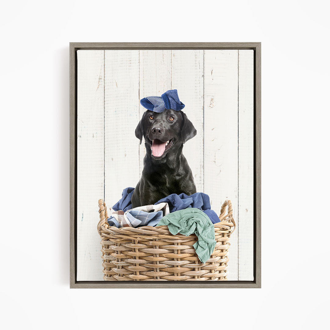 A black dog wearing a blue hat sits in a wicker basket filled with clothes.
