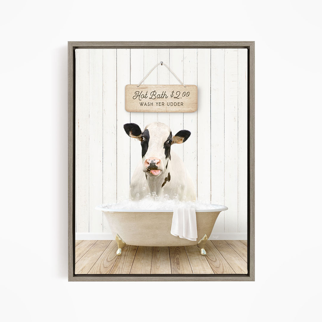 A black and white cow is sitting in a white bathtub filled with water, with a wooden sign above the tub that reads "Hot Bath $2.00 WASH YER UUDDER".