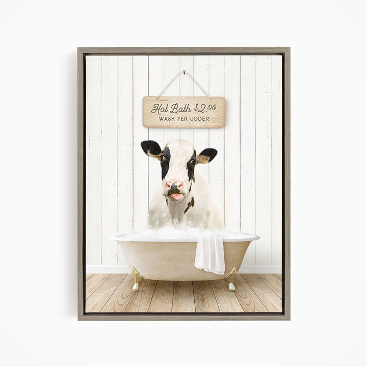 A black and white cow is sitting in a white bathtub filled with water, with a wooden sign above the tub that reads "Hot Bath $2.00 WASH YER UUDDER".