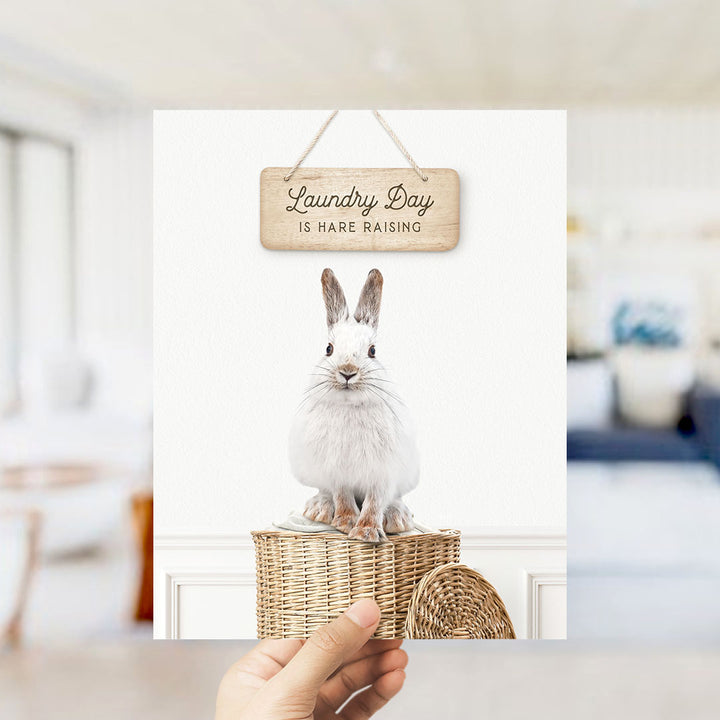 A hand holding a sign that says "Laundry Day is Hare Raising" above a picture of a white rabbit sitting in a wicker basket.