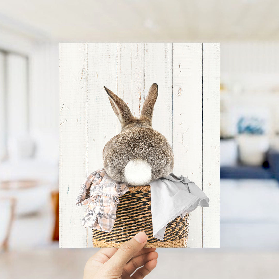 A person's hand is holding up a picture of a rabbit sitting in a basket, with a blurred background suggesting an indoor setting.