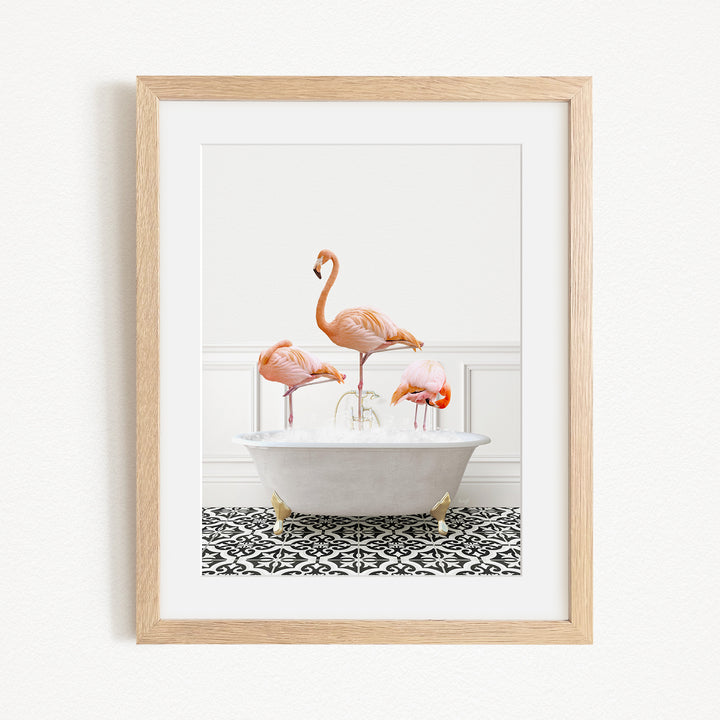 A framed image depicting three flamingos standing in a white bathtub filled with water, set against a white wall and black and white patterned floor.