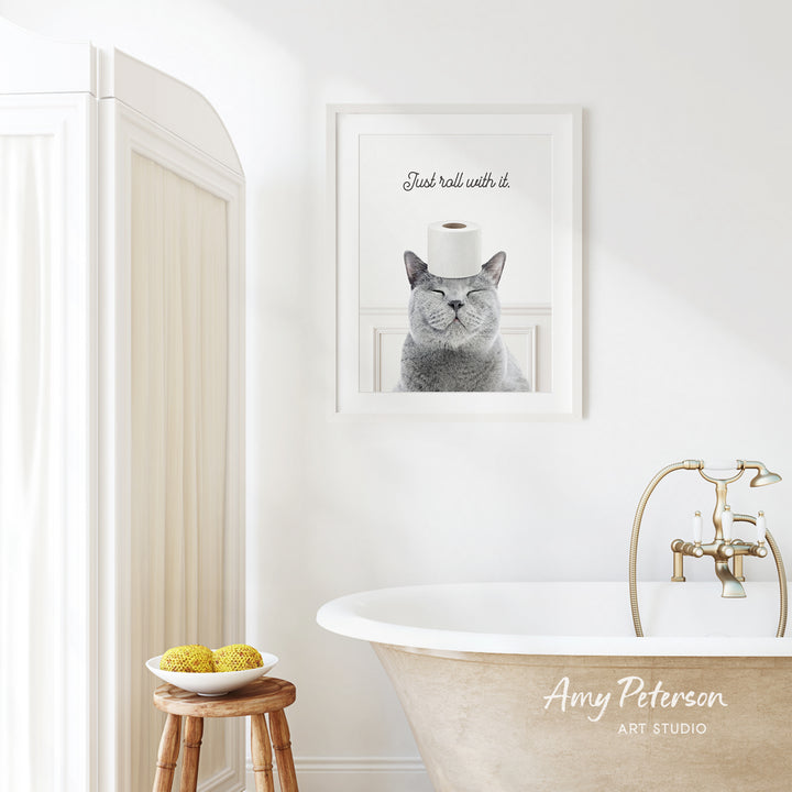 A framed image of a cat with a roll of toilet paper on its head is displayed on the wall above a white bathtub in a bathroom.