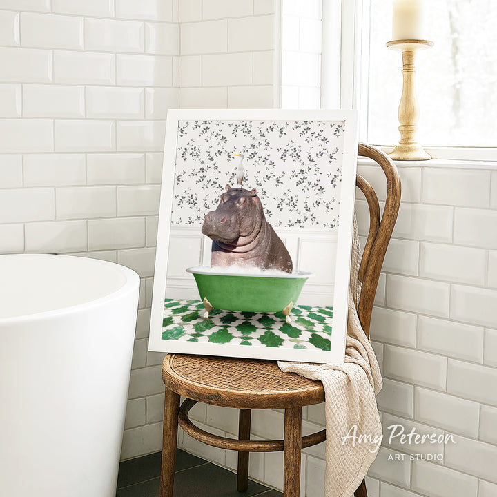 A framed image of a hippopotamus sitting in a green bathtub, surrounded by a patterned tile design, is displayed on a wooden chair in a bathroom setting.