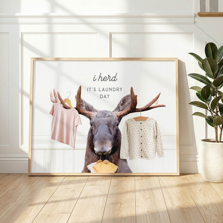 A framed poster with a moose holding clothes, accompanied by the text "i herd, it's laundry day"