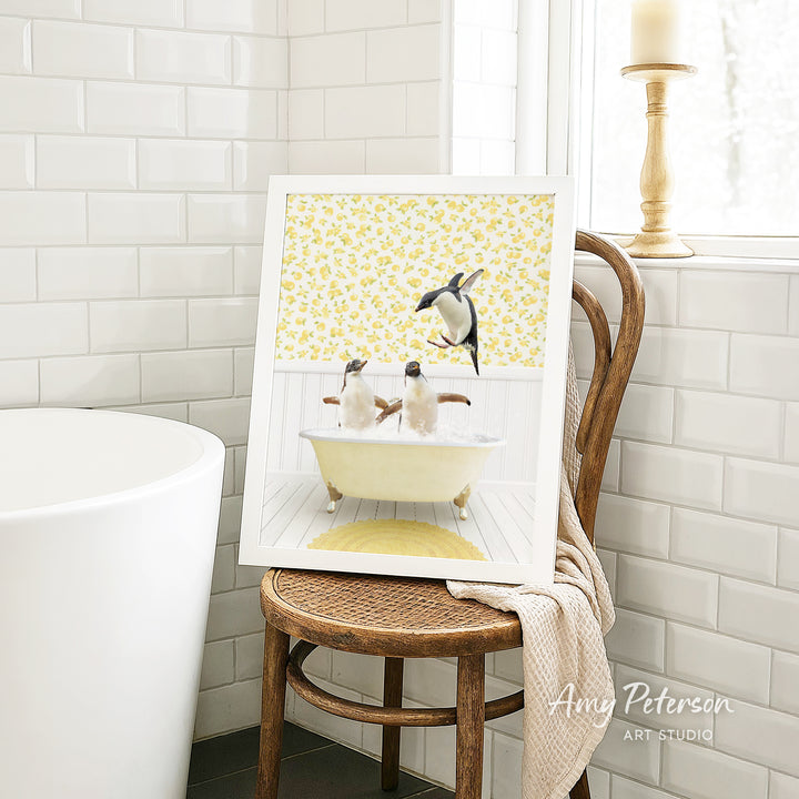 A framed image of two penguins sitting in a bathtub, surrounded by a yellow floral pattern, is displayed on a wooden chair in a bathroom setting.