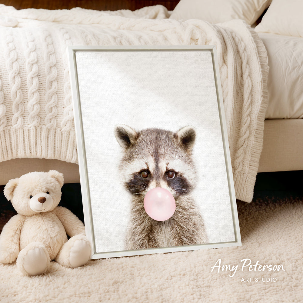 A framed image of a raccoon blowing a bubble with a pink bubble gum bubble, accompanied by a teddy bear on a bed.