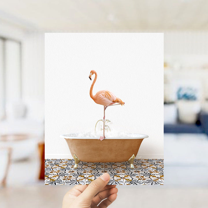 A hand holds up a framed image of a pink flamingo standing in a bathtub filled with water and bubbles.