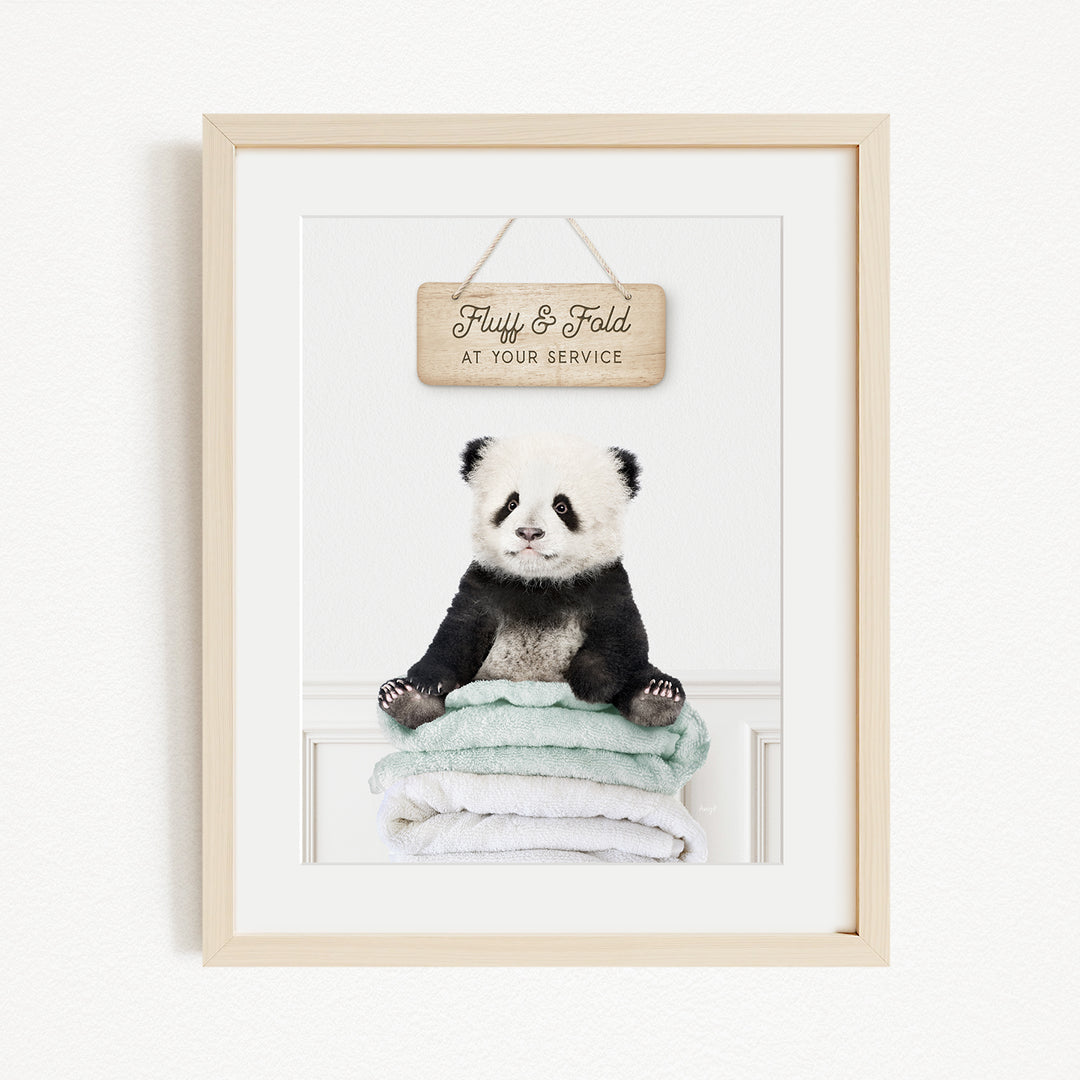 A framed image of a panda bear sitting on a stack of towels, with a wooden sign above that says "Fluffy & Fold at your service".