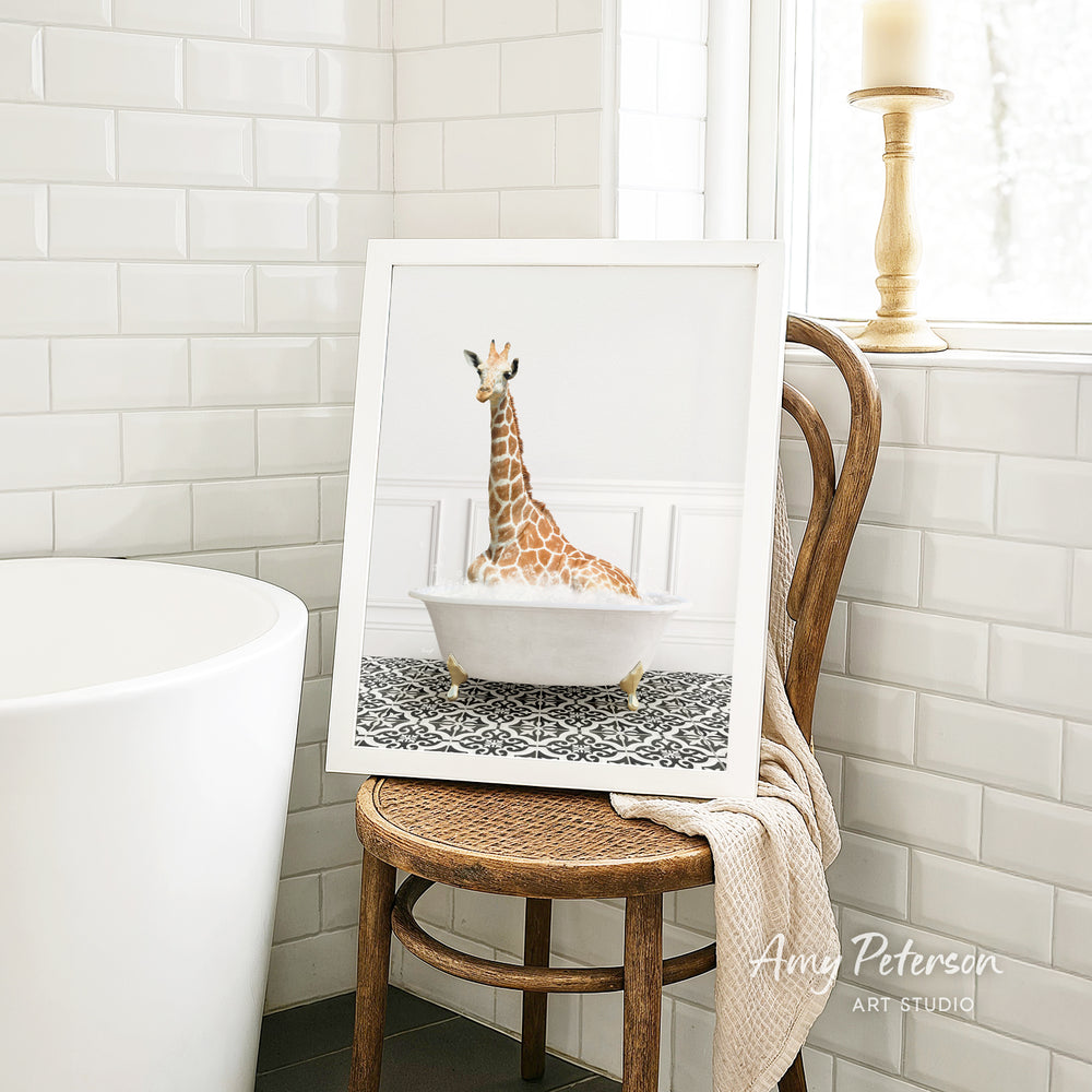 A framed image of a giraffe sitting in a bathtub is displayed on a wooden chair in a bathroom setting.