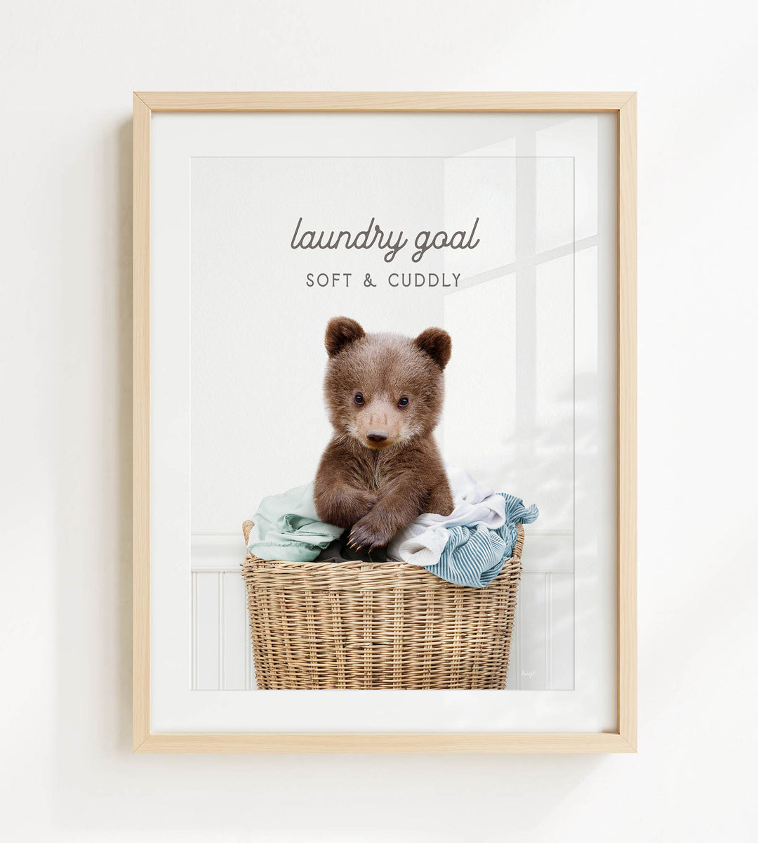 A cute brown bear sitting in a wicker laundry basket filled with clothes, with the text "laundry goal: soft & cuddly" displayed above the bear.