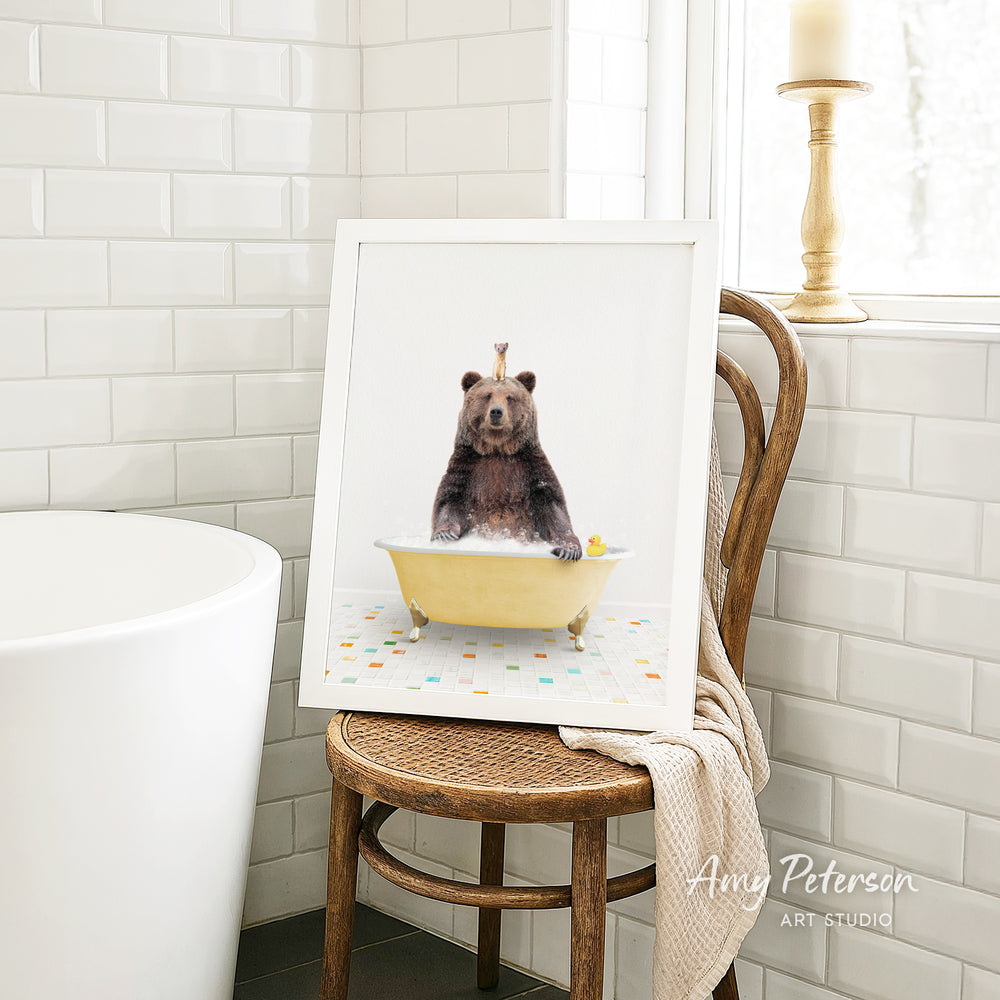A framed image of a bear sitting in a bathtub, with a yellow bathtub and a towel draped over the back of a wooden chair.