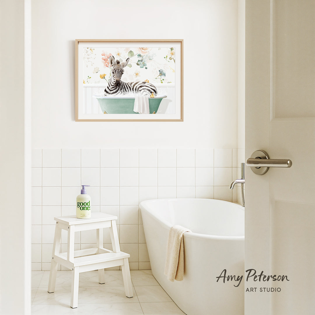 A framed painting of a zebra sitting in a bathtub is hanging on a white tiled wall above a white bathtub in a bathroom.