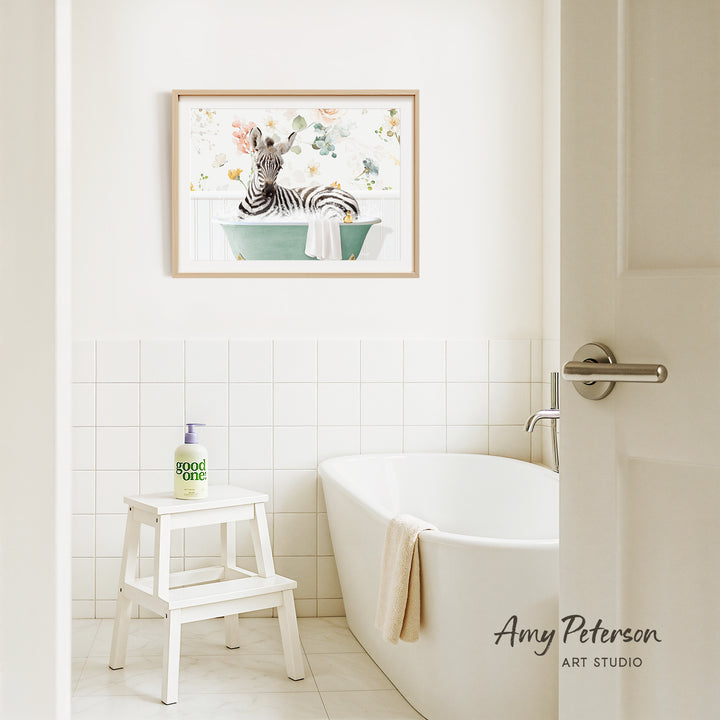 A framed painting of a zebra sitting in a bathtub is hanging on a white tiled wall above a white bathtub in a bathroom.