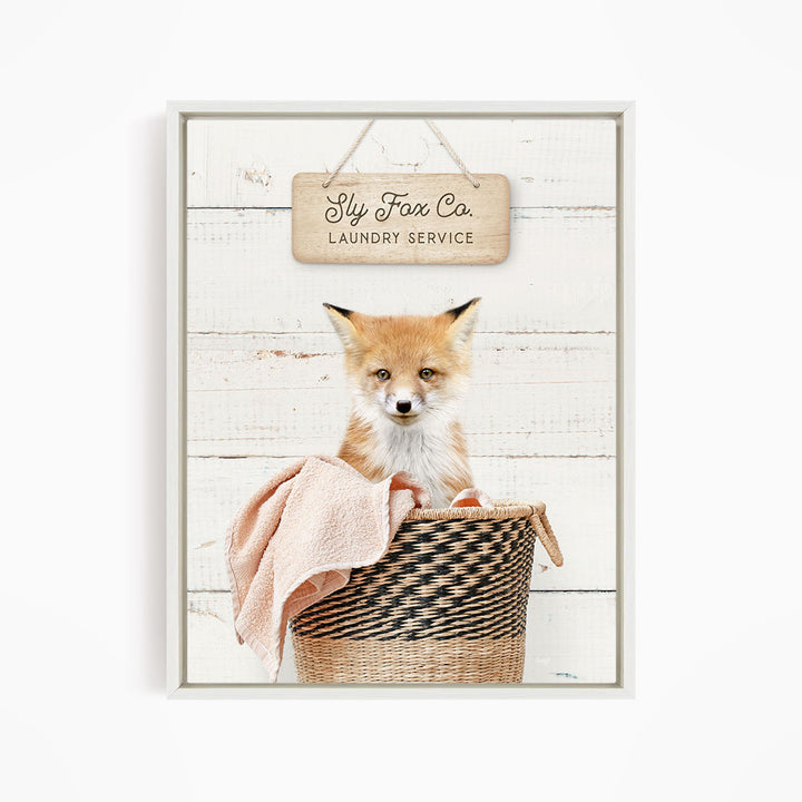 A young fox sits in a woven basket, with a sign above that reads "Laundry Service" in a rustic, wooden frame.