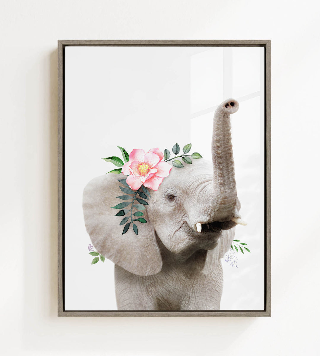 A framed image of an elephant with a pink flower and green leaves adorning its head.