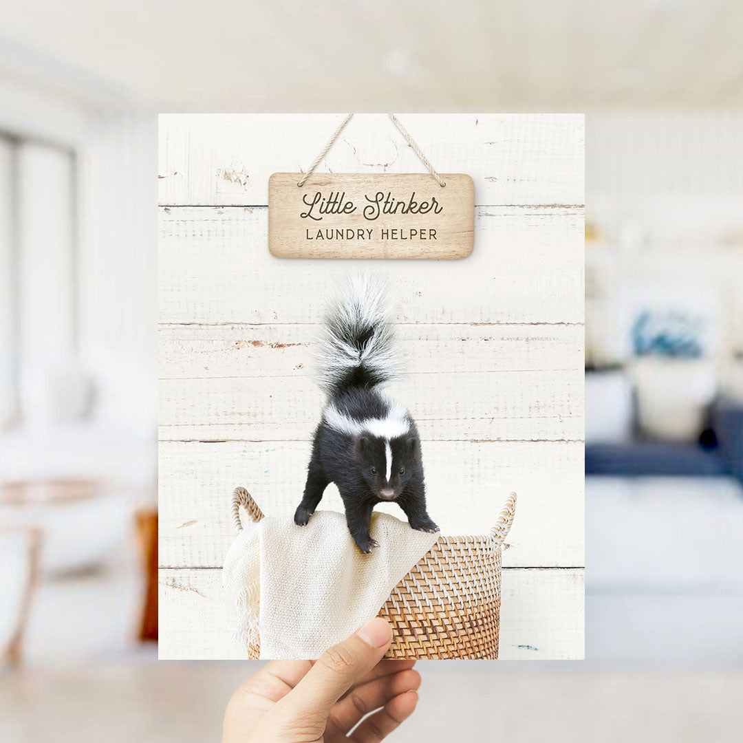 A hand holds up a framed image of a skunk standing on a woven basket, with a wooden sign above it that reads "Little Stinker Laundry Helper".