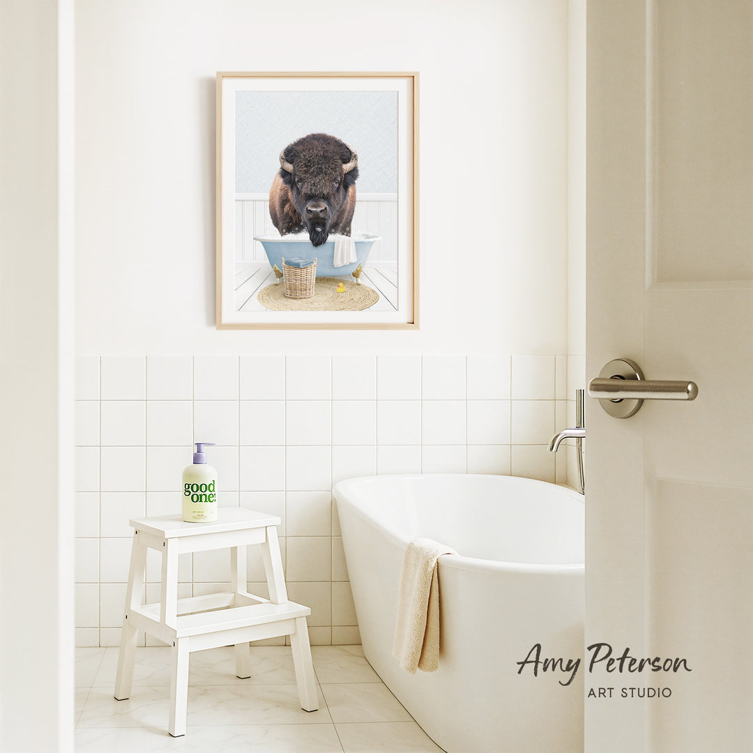a bathroom with a framed picture of a buffalo in a bathtub, a white bathtub, a white stool, and a bottle of soap on a white stool.