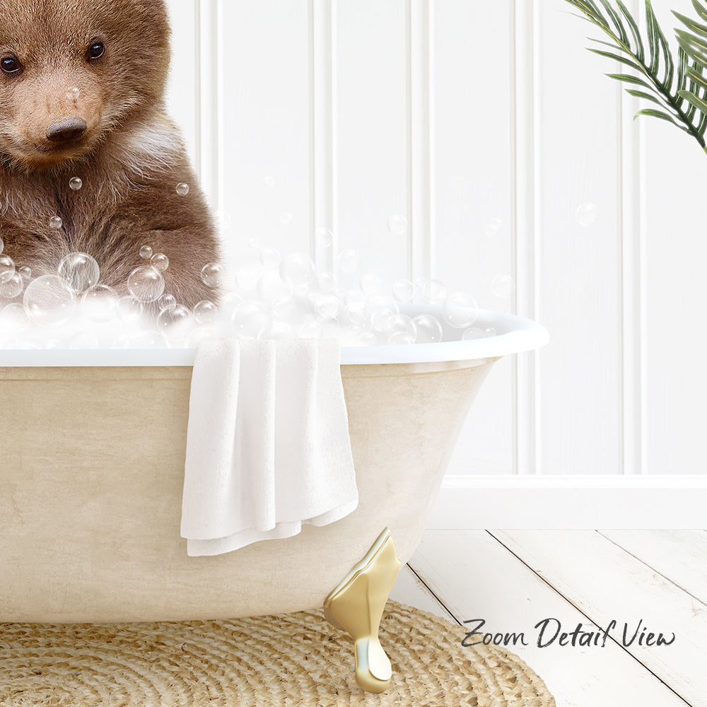 A cute brown bear cub sitting in a white bathtub filled with bubbles, with a towel and a plant nearby.