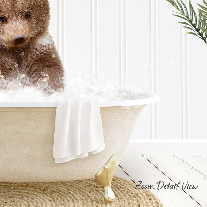 A cute brown bear cub sitting in a white bathtub filled with bubbles, with a towel and a plant nearby.