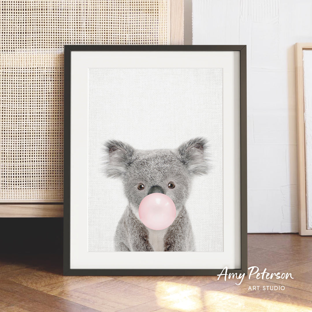 A framed image of a koala bear blowing a bubble with a pink bubblegum bubble.