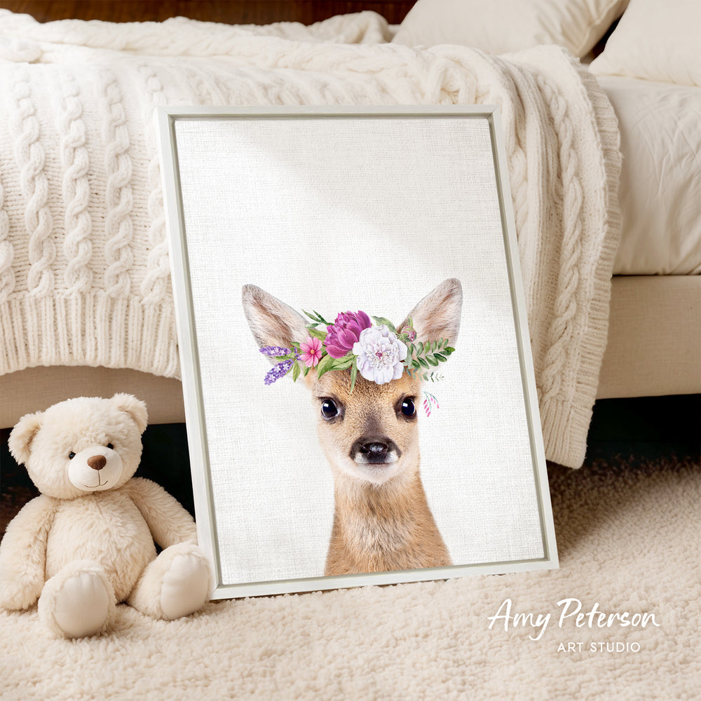 A framed print of a deer wearing a flower crown is displayed on a white bedspread, accompanied by a teddy bear on the floor.