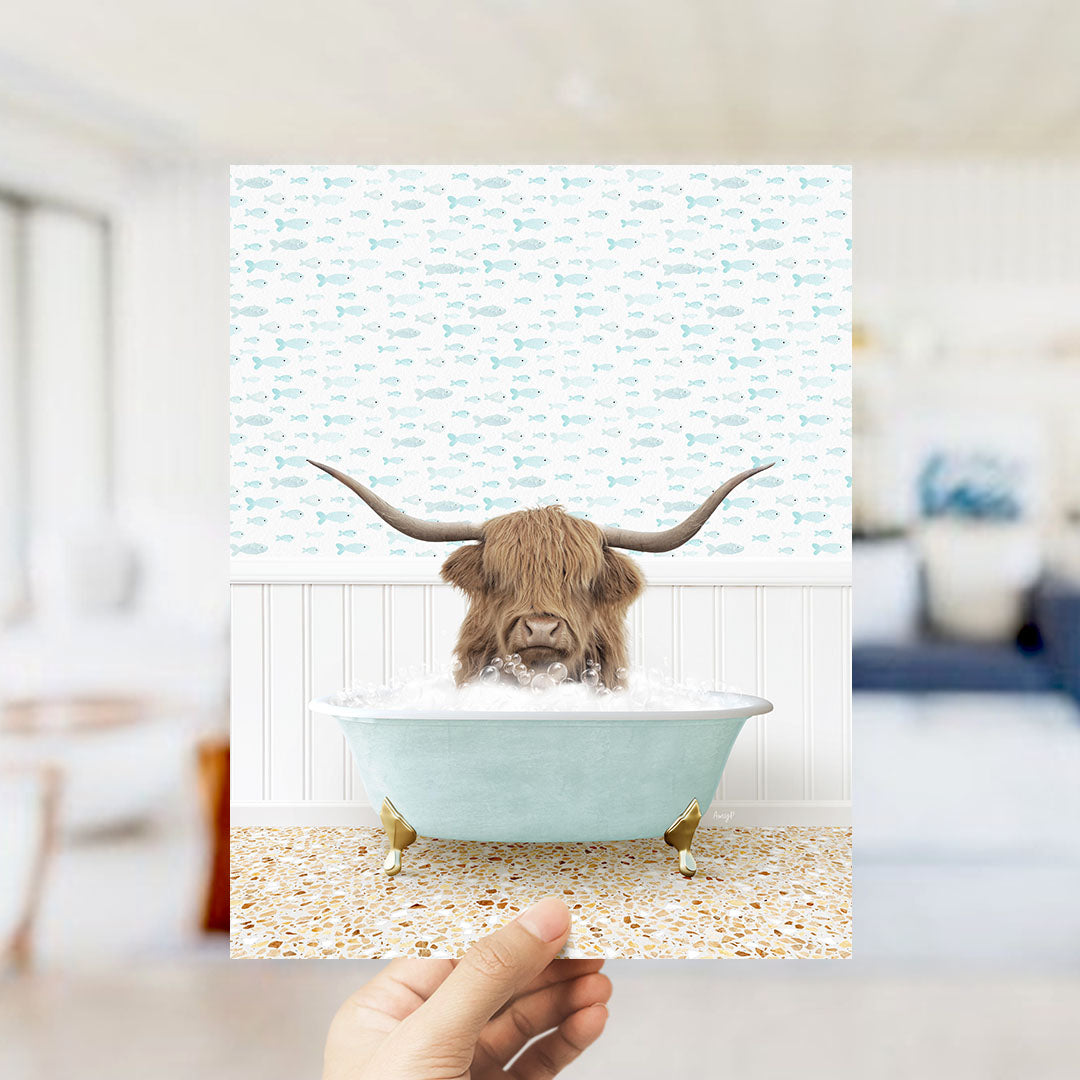 A hand holds up a framed image depicting a Highland cow with large curved horns sitting in a light blue bathtub filled with bubbles.