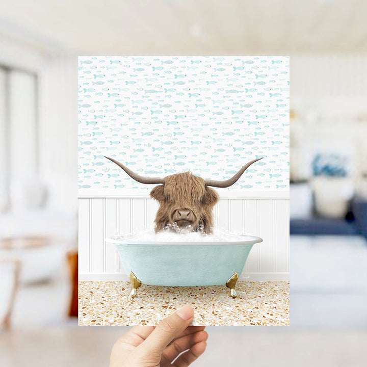 A hand holds up a framed image depicting a Highland cow with large curved horns sitting in a light blue bathtub filled with bubbles.