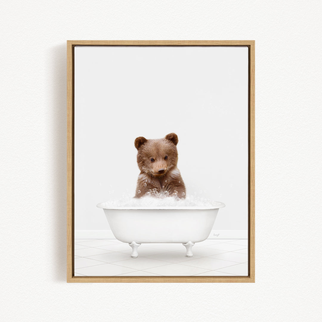 A cute brown bear sitting in a white bathtub filled with bubbles, looking directly at the camera.