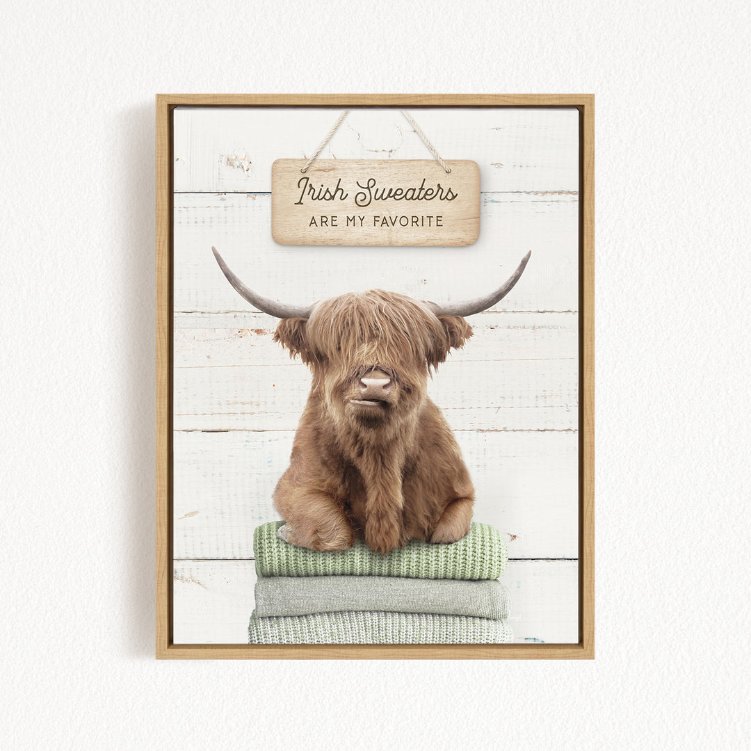 A brown Highland cow with long horns is sitting on a stack of green blankets, with a wooden sign above that says "Irish Sweaters are my favorite".