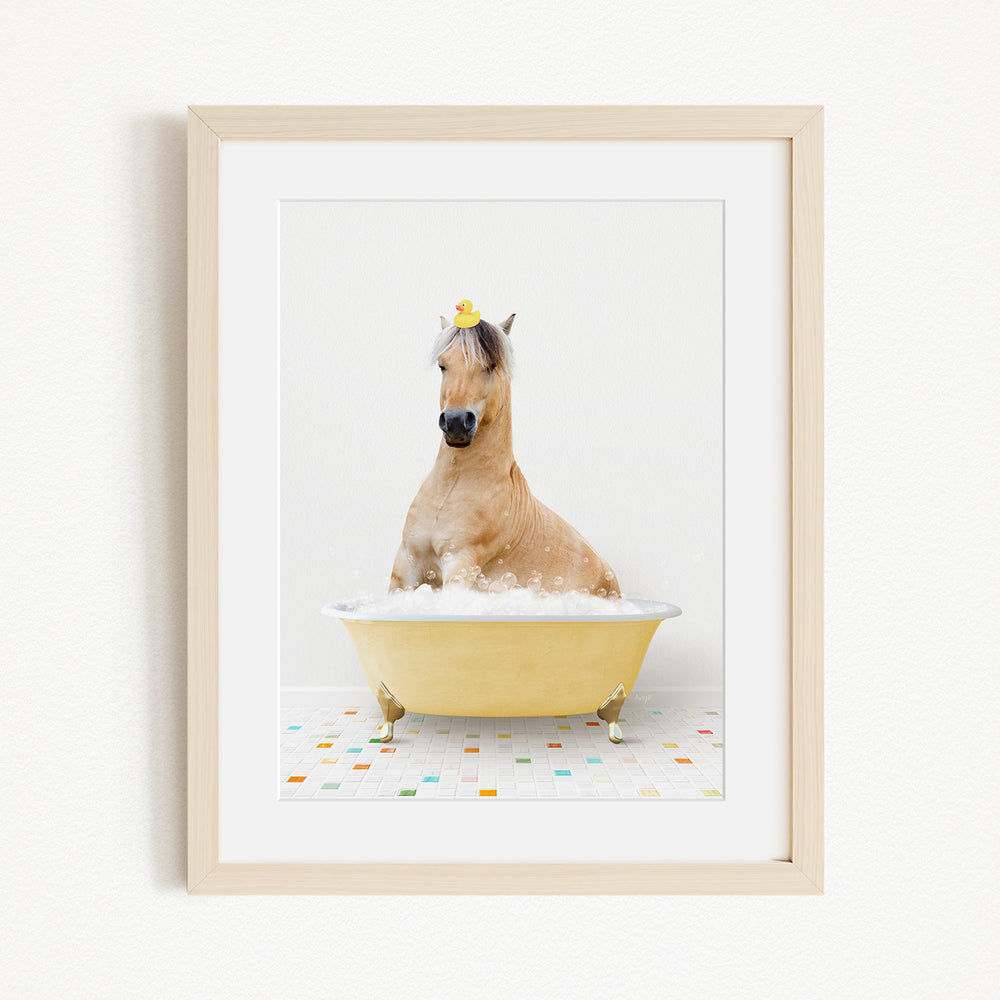 A light brown horse is sitting in a yellow bathtub filled with bubbles, with a yellow rubber duck perched on its head.