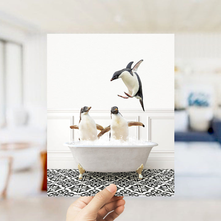 A hand holds up a framed photograph of two penguins standing in a bathtub filled with water, with one penguin flying out of the tub.