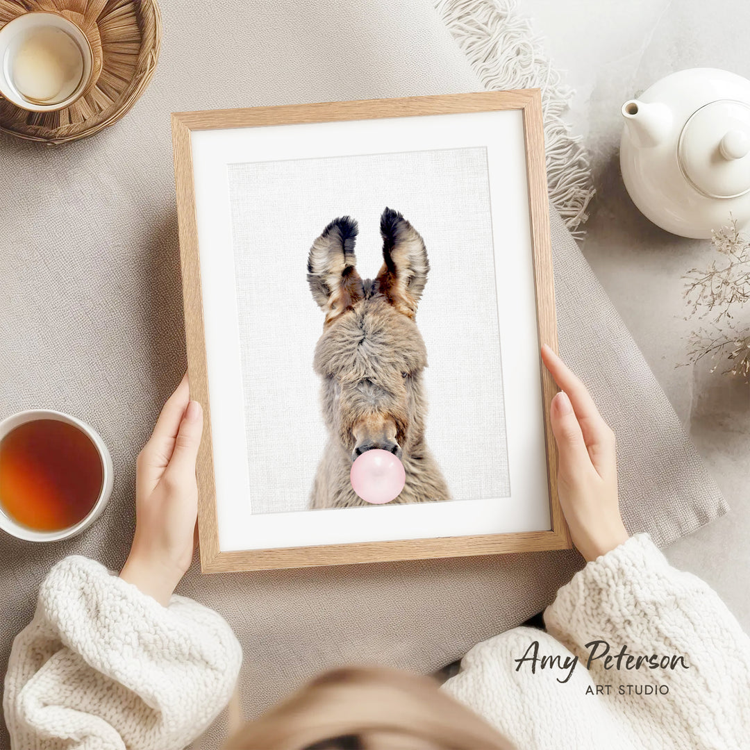A person is holding a framed picture of a donkey with a pink bubble gum bubble in its mouth, surrounded by various objects such as a teapot, a cup, and a bowl.