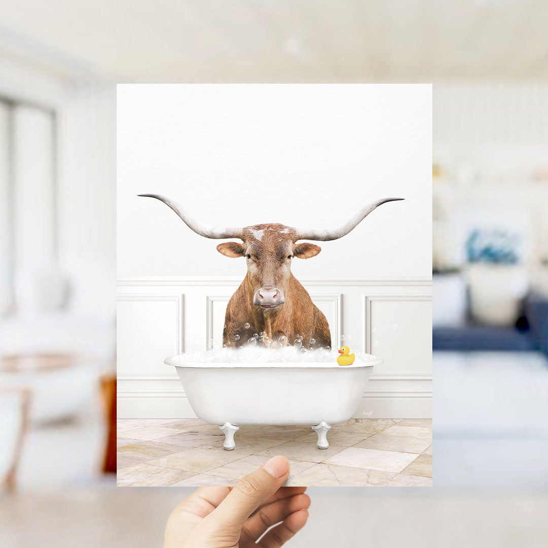 A brown cow with long horns is sitting in a white bathtub filled with bubbles, with a yellow rubber duck floating in the water.