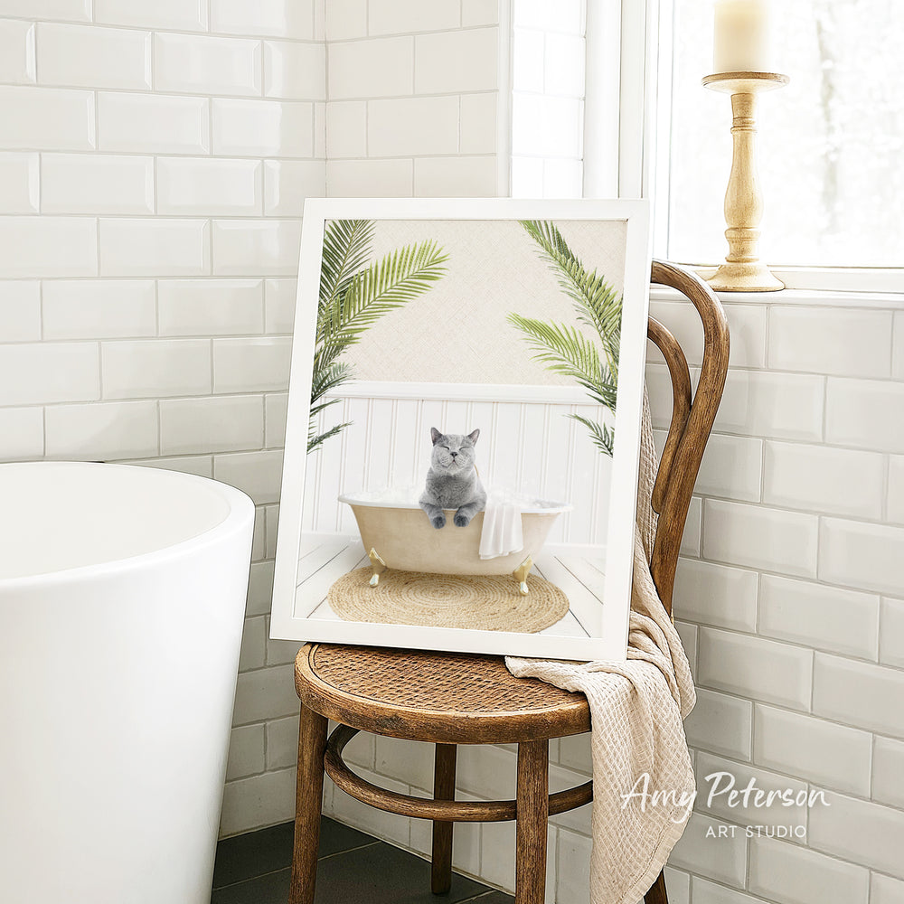 A framed image of a gray cat sitting in a bathtub, surrounded by green plants, is displayed on a wooden chair in a bathroom setting.