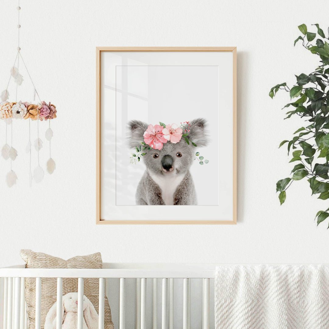 A framed picture of a koala bear wearing a flower crown is hanging on a white wall in a nursery.