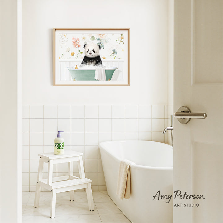 A framed painting of a panda sitting in a bathtub is hanging on a white tiled wall above a white bathtub in a bathroom.