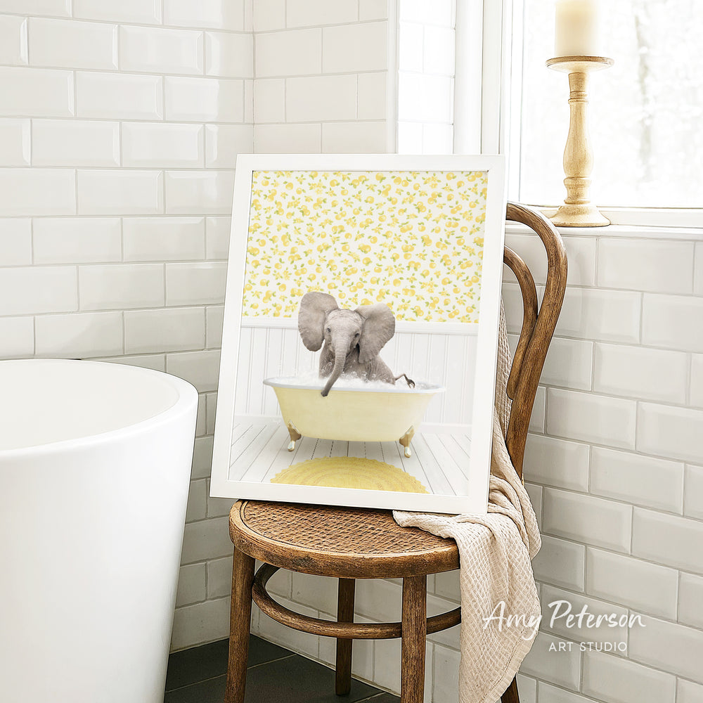 A framed image of an elephant sitting in a bathtub, surrounded by yellow flowers, is displayed on a wooden chair in a bathroom setting.