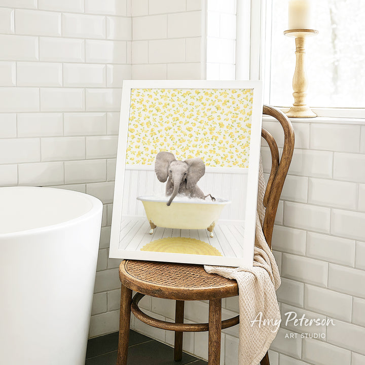 A framed image of an elephant sitting in a bathtub, surrounded by yellow flowers, is displayed on a wooden chair in a bathroom setting.
