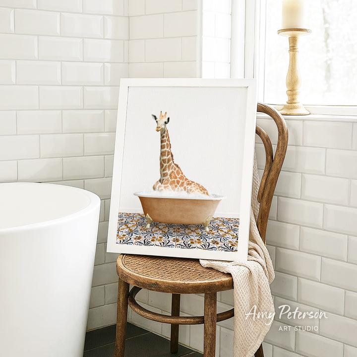 A framed image of a giraffe sitting in a bathtub is displayed on a wooden chair in a bathroom setting.