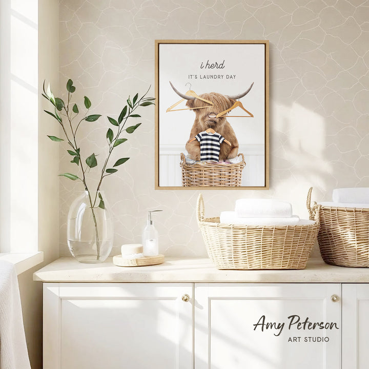 a framed poster of a bull with a laundry basket, accompanied by a vase of leaves and a bottle of soap on a white cabinet.
