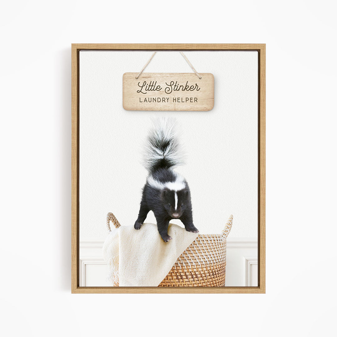 A framed image of a black and white skunk standing on a woven basket, with a wooden sign above it that reads "Little Stinker Laundry Helper".